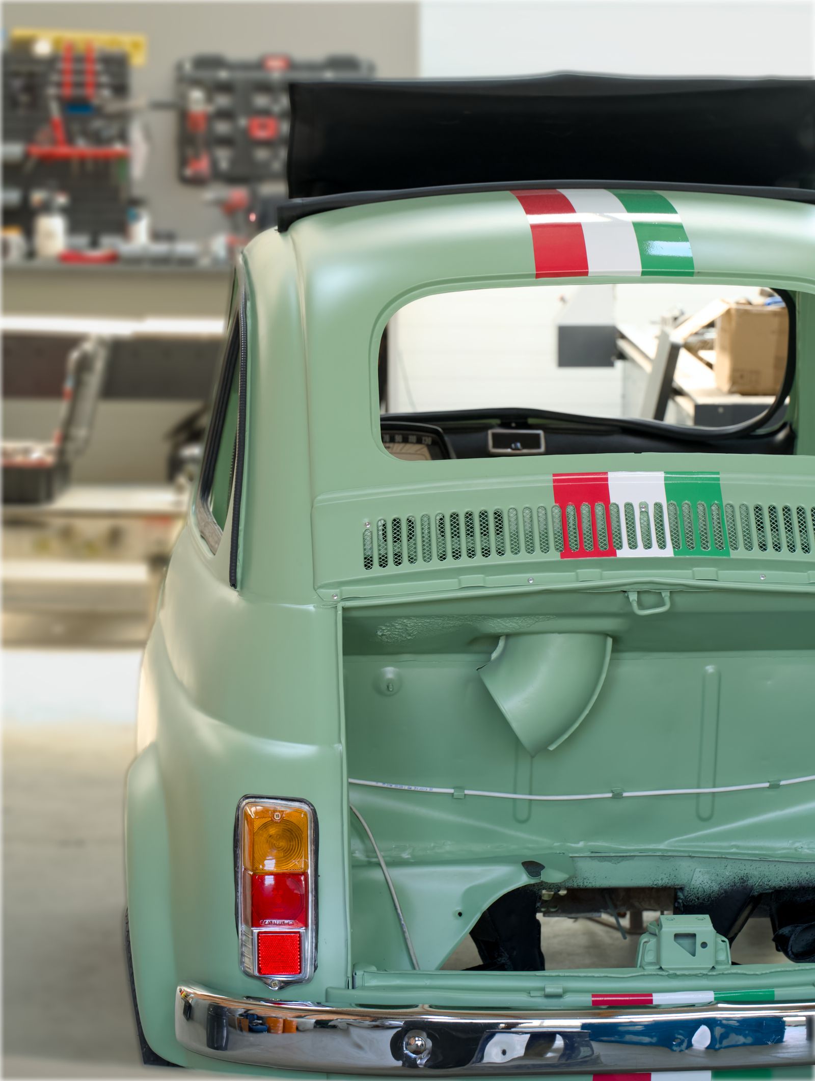 Rear three-quarter view of a freshly restored mint-green vintage city car with red-white-green racing stripes, glossy new paint and chrome bumper, parked inside a bright workshop.