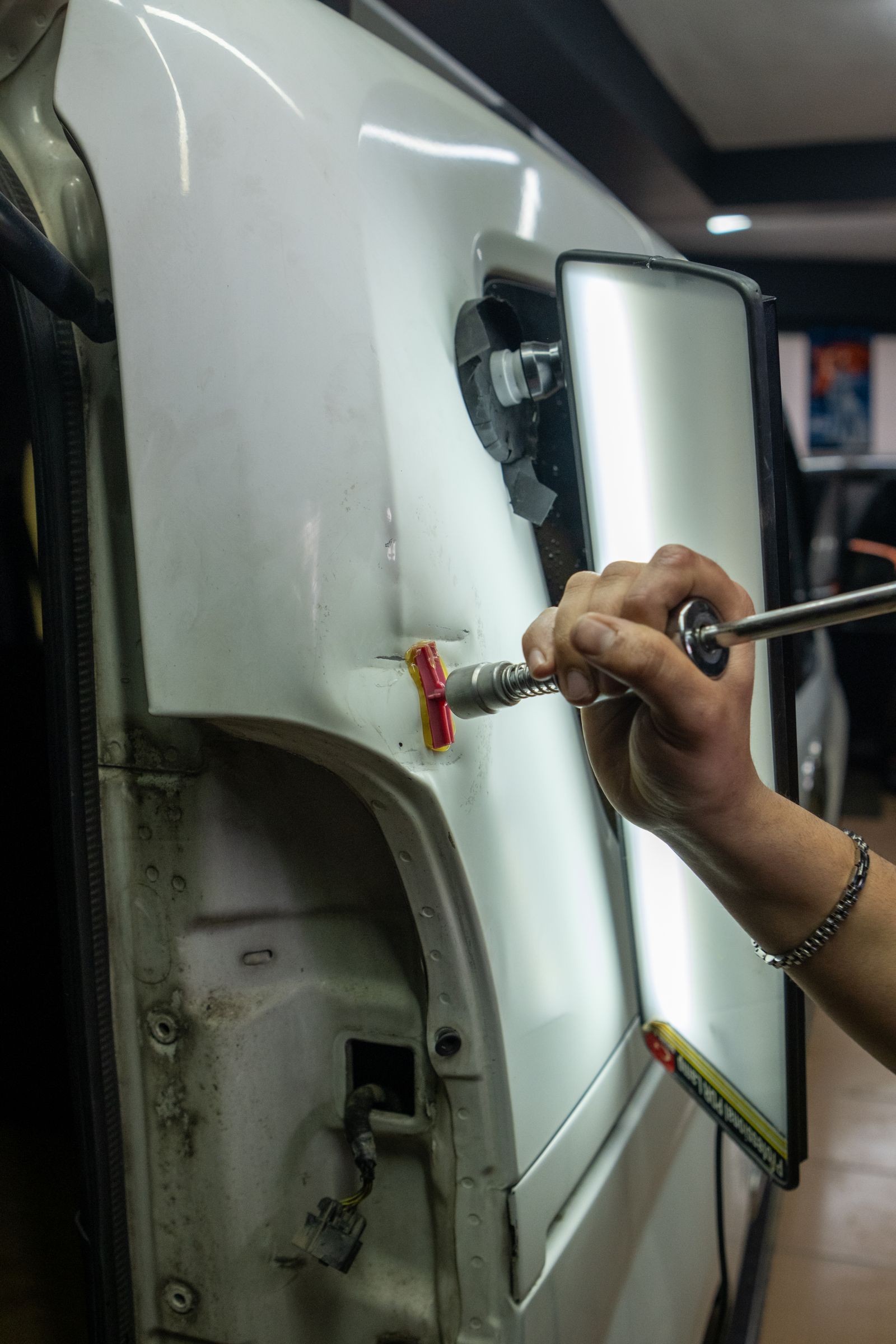 A hand using a slide-hammer glue-pull dent-removal tool on a white car body panel, with a striped LED reflector board held against the panel to read the surface.
