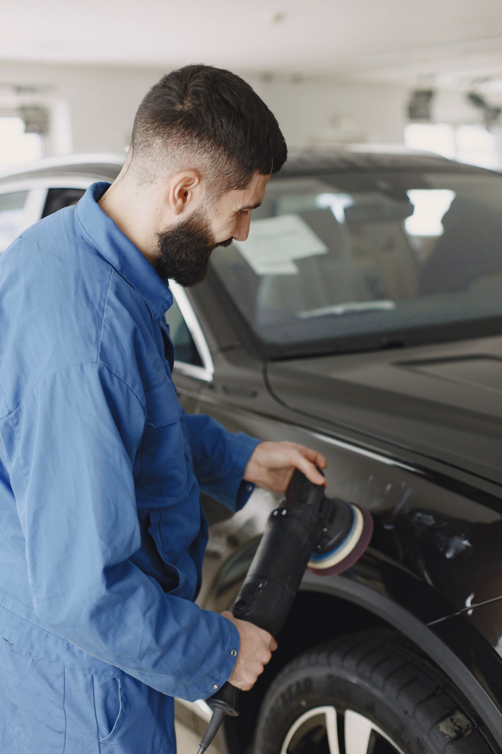 A technician in blue overalls using a dual-action machine polisher on the glossy black panel of a car inside a workshop, with a light-coloured car out of focus behind.