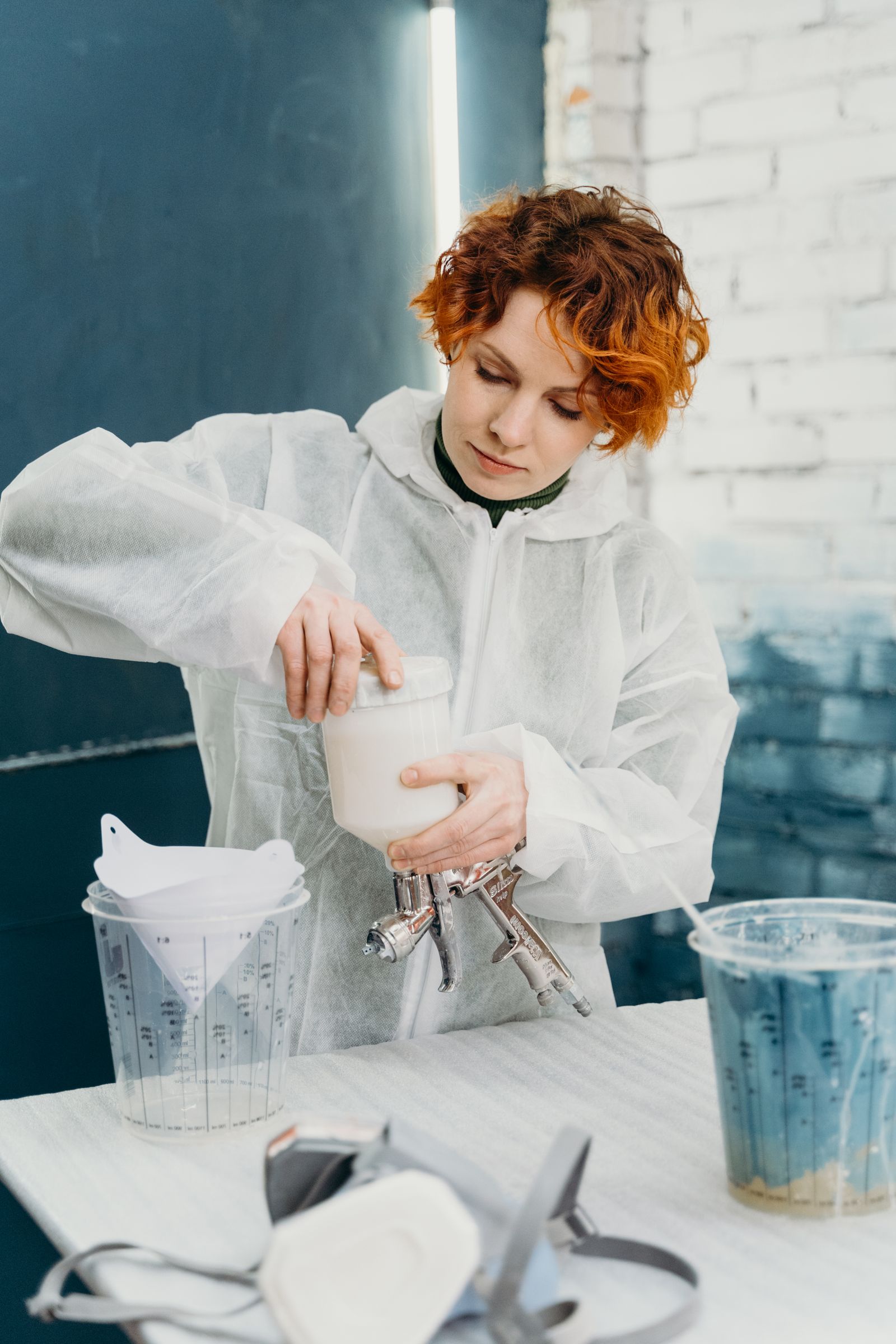 A person in a white protective suit standing at a bench mixing and decanting paint into a spray-gun cup, with measuring jugs and a respirator on the bench.