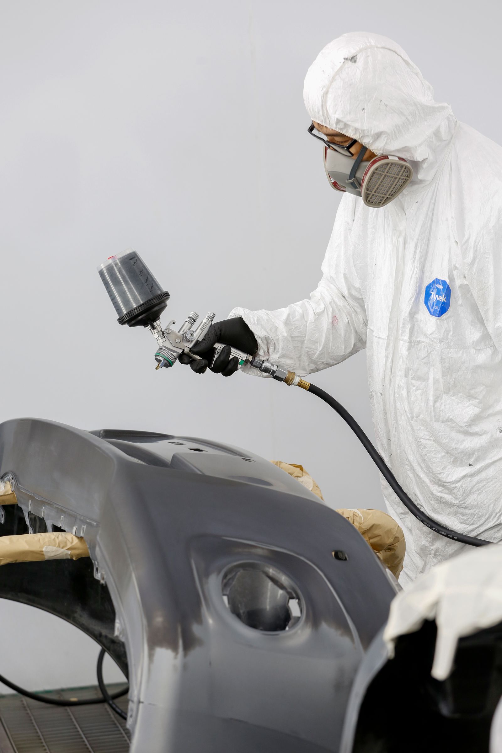 A painter in a white hooded coverall and respirator spraying grey primer onto a car bumper clamped on a stand in a spray booth, with masking paper visible.